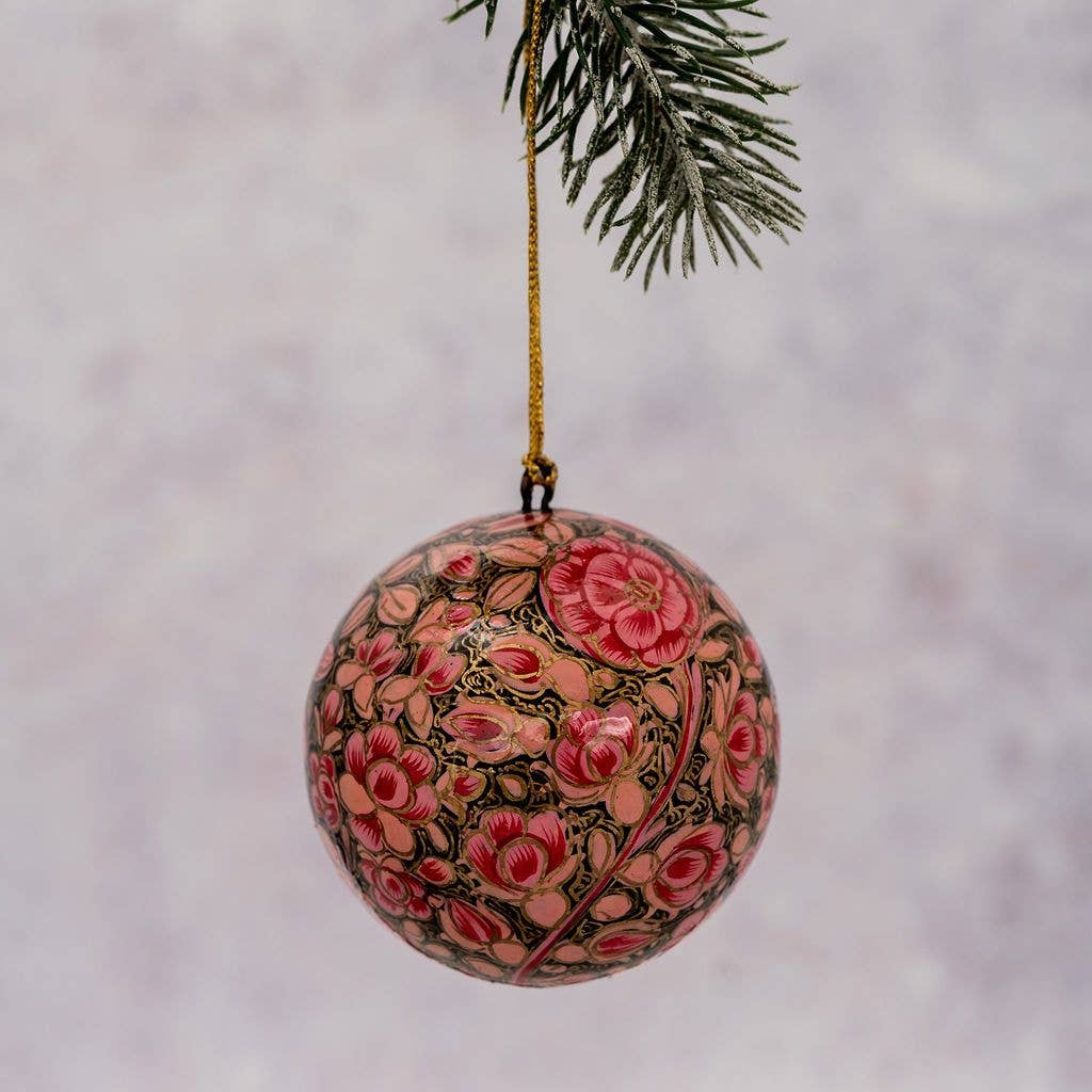 A hand-painted wooden Christmas ornament with a pink and red floral pattern on a dark background, hanging from a gold string next to a pine branch.