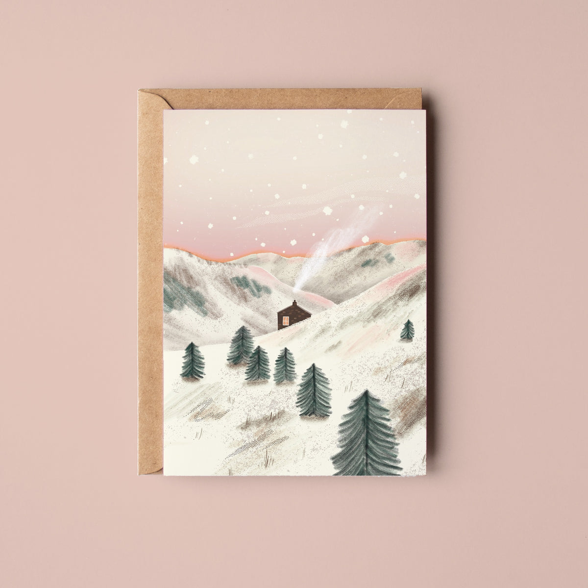 A greeting card with a kraft envelope, featuring an illustration of a snowy mountain landscape with a small cabin and evergreen trees, set against a solid dusty pink background. Snowy Bothy Greetings Card by Rachel Akerman at The Country Crib