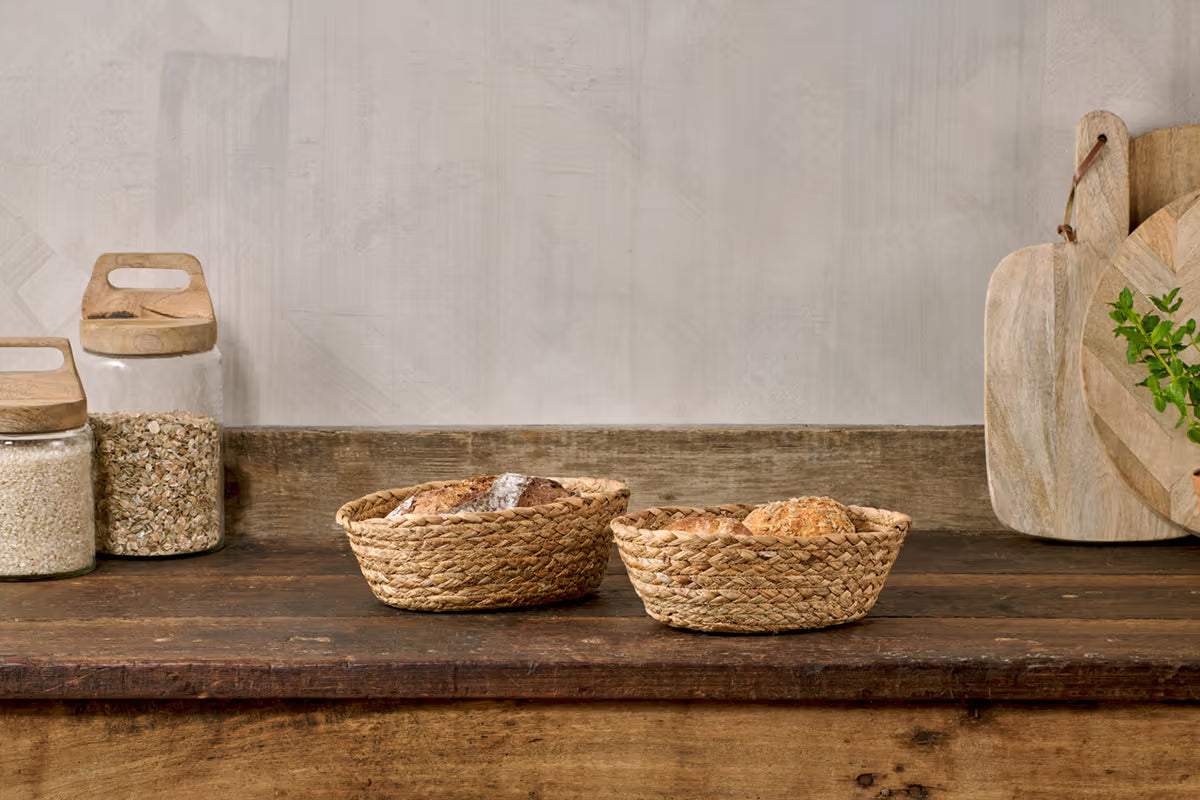 Two woven baskets on a wooden shelf with jars and a cutting board in the background.