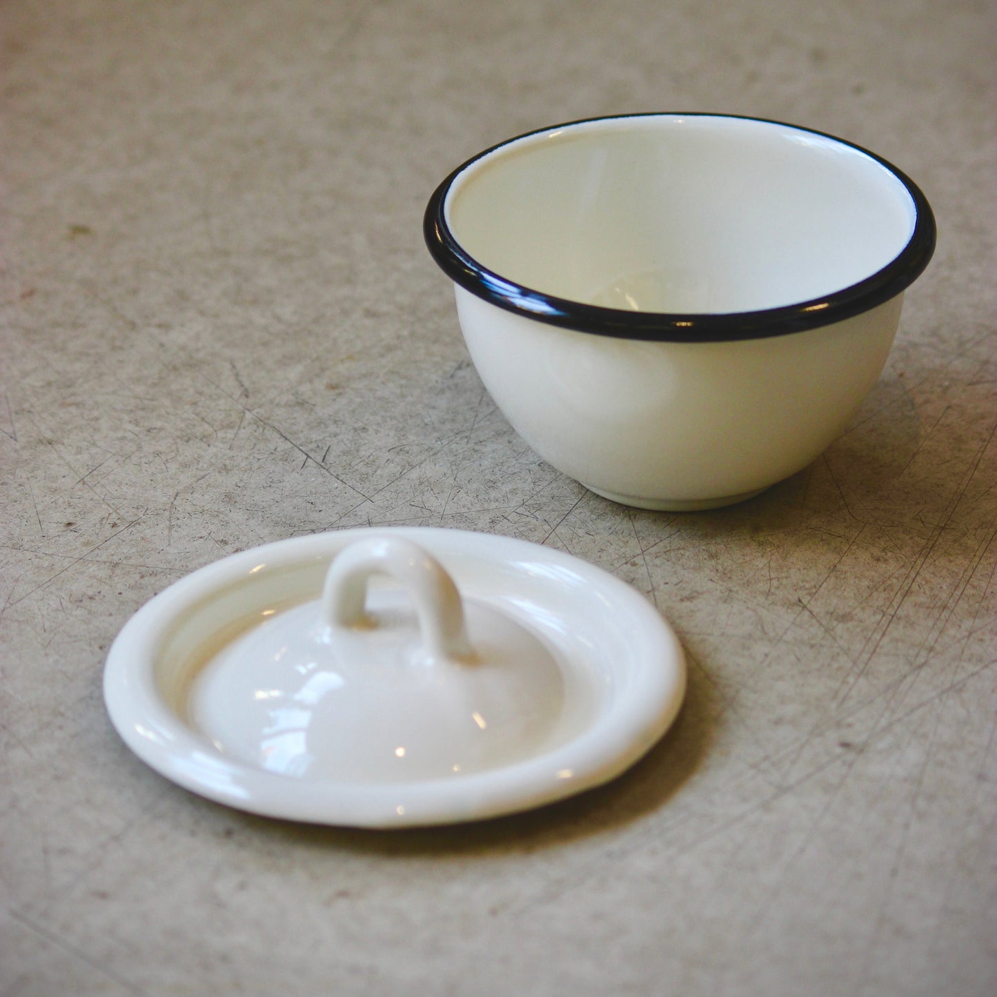 White enamel bowl with black rim and matching lid on a beige surface