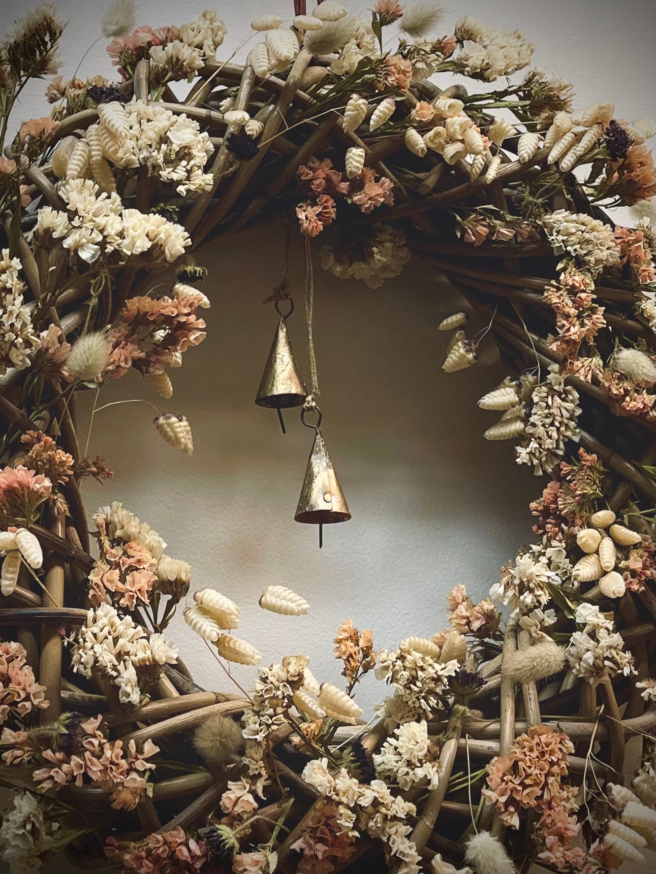 Decorative wreath with dried flowers and a hanging bell against a neutral background