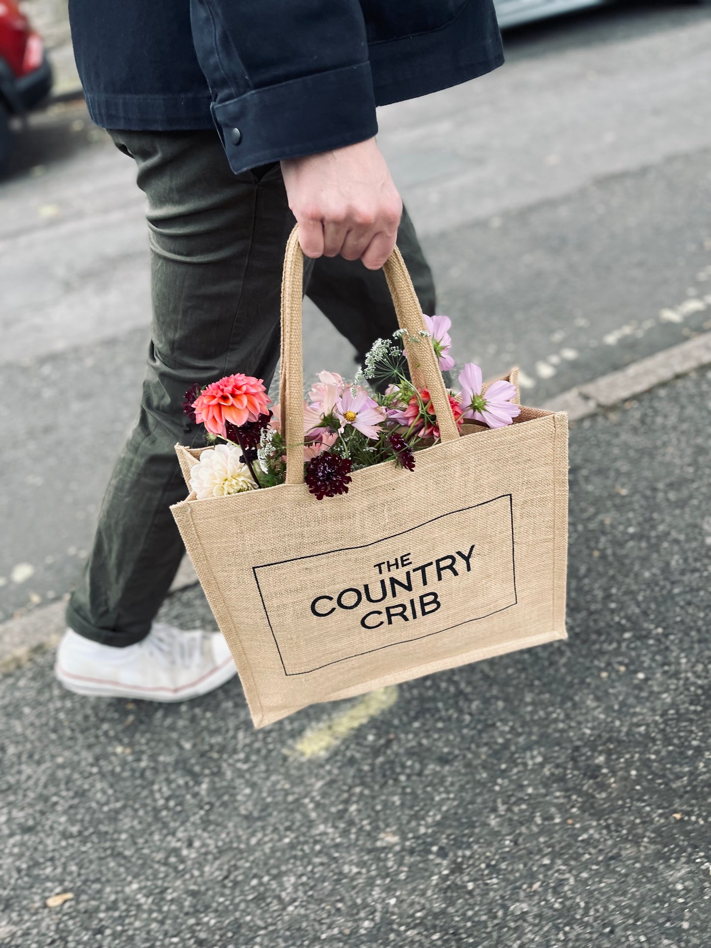 Person holding a tote bag with flowers labeled 'The Country Crib' on a street.