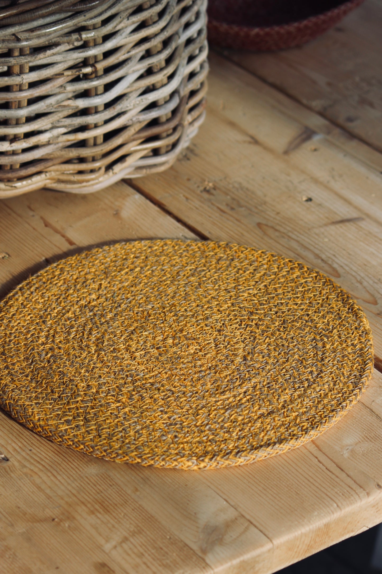 A faire trade round woven jute placemat in a golden yellow mustard, made in Bangladesh, displayed on a light wood table next to a wicker basket.