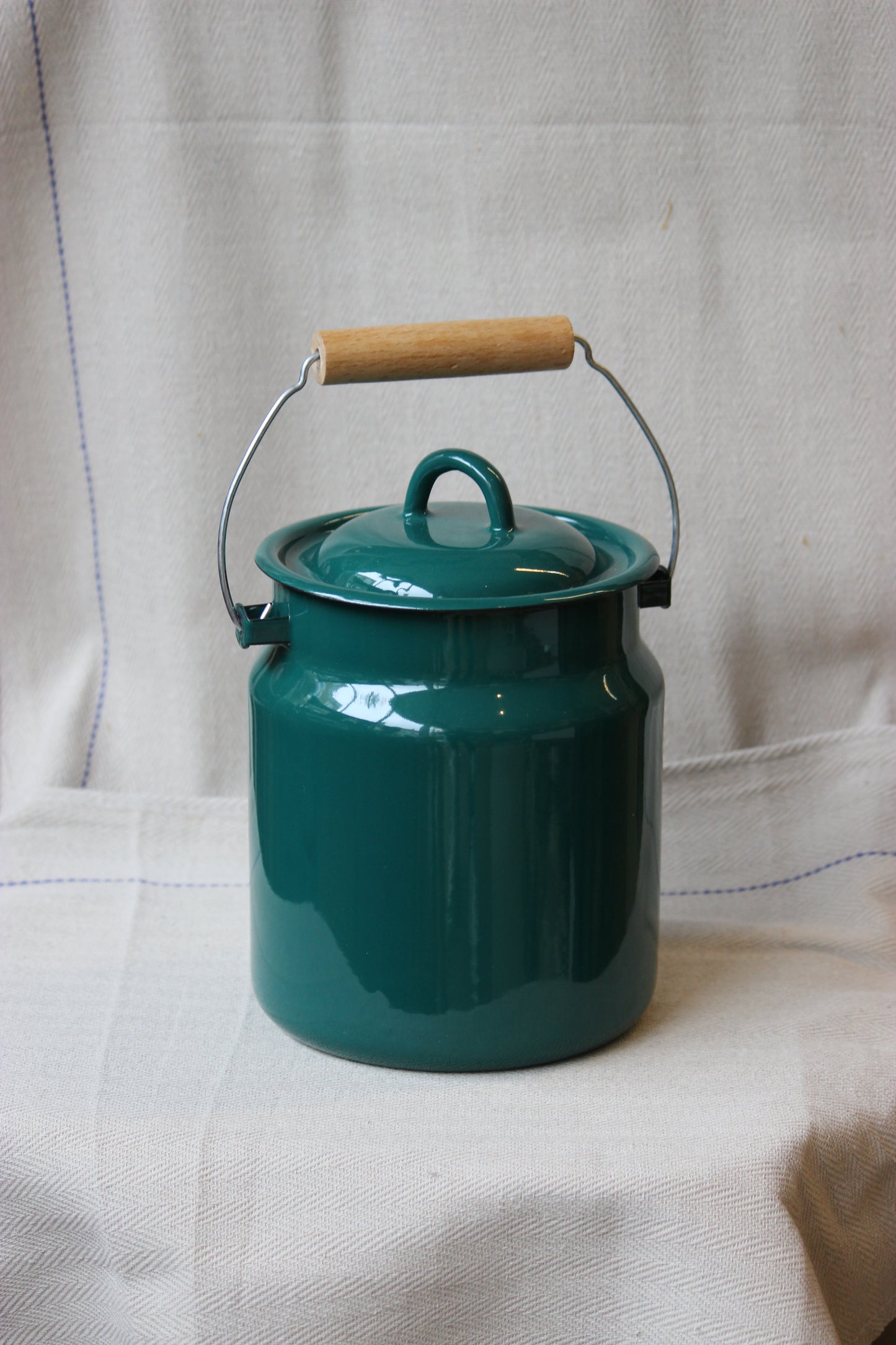 Teal enamelware storage jar made in Ukraine with wooden handle on a white fabric background