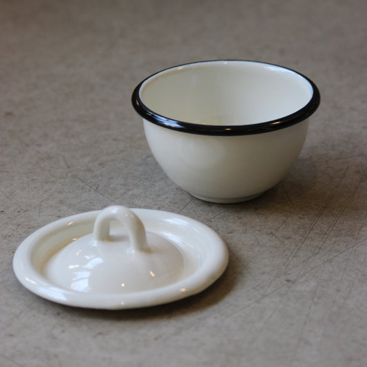 White enamel bowl with black rim and matching lid on a beige surface