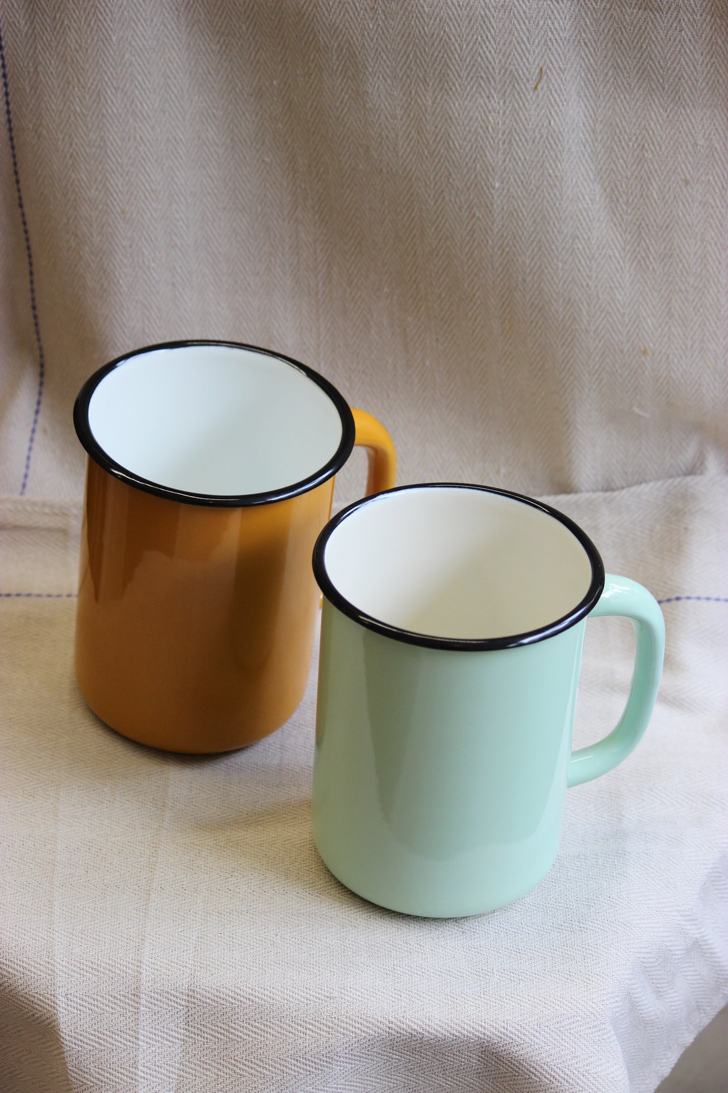 Two enamel jugs, one green and one mustard, on a textured fabric surface.