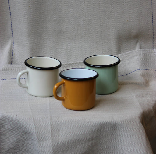 Three enamel mugs in white, orange, and green on a textured fabric background