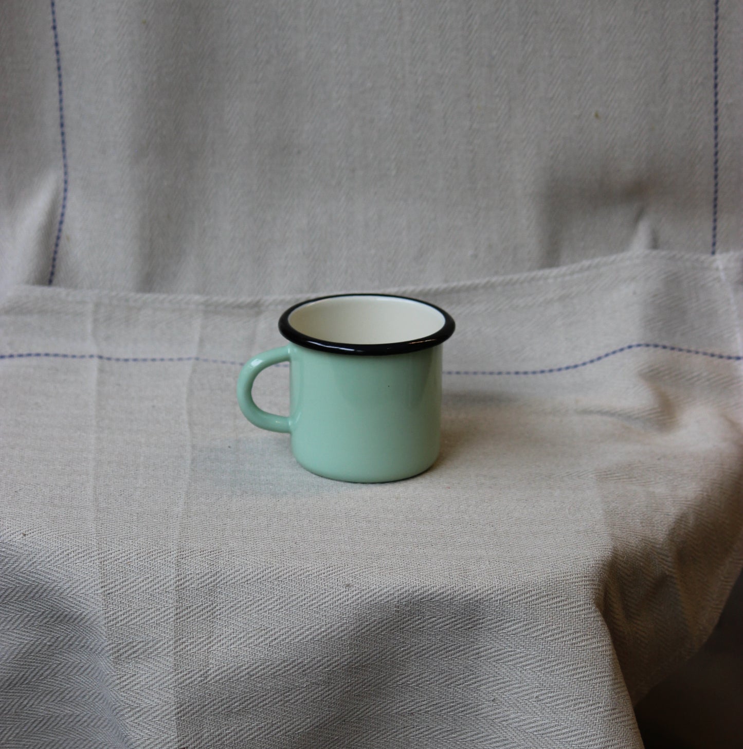 Small green enamel mug with black rim on a textured fabric background
