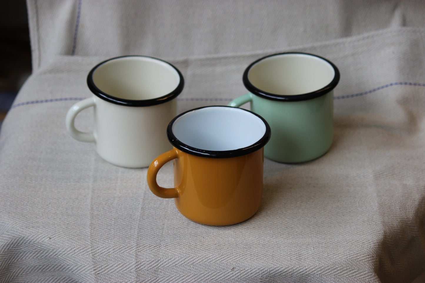 Three enamel mugs in white, green, and orange on a textured fabric surface.