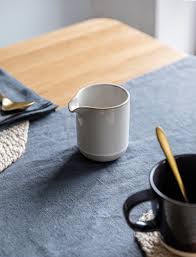 White ceramic pitcher on a blue tablecloth with a black mug and wooden spoon.