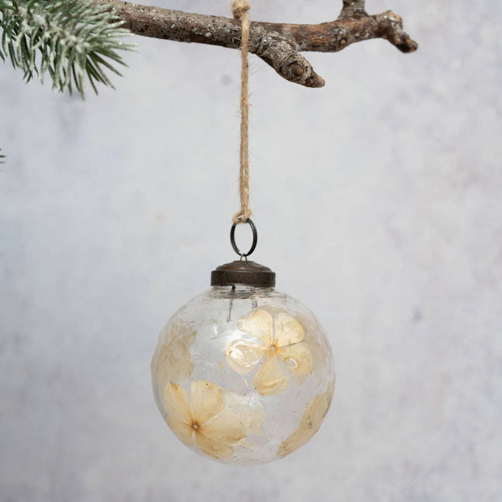 A clear glass Christmas ornament hanging from a rustic branch with a jute string. The ornament is transparent and contains dried cream-colored flowers pressed inside, with a dark metal cap and ring at the top.