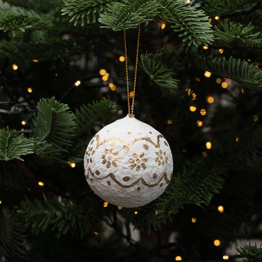 A textured cotton paper mache white Christmas bauble ornament with gold glitter floral patterns, hanging from a gold string on a green Christmas tree with warm bokeh lights.