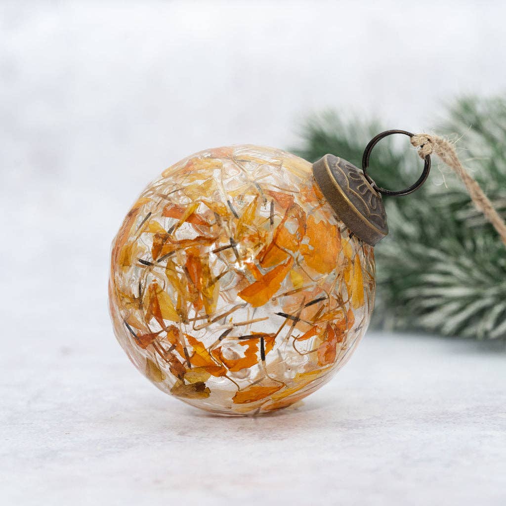A clear glass Christmas ornament ball filled with dried orange and yellow botanical pieces, resting on a light surface with blurred greenery in the background.