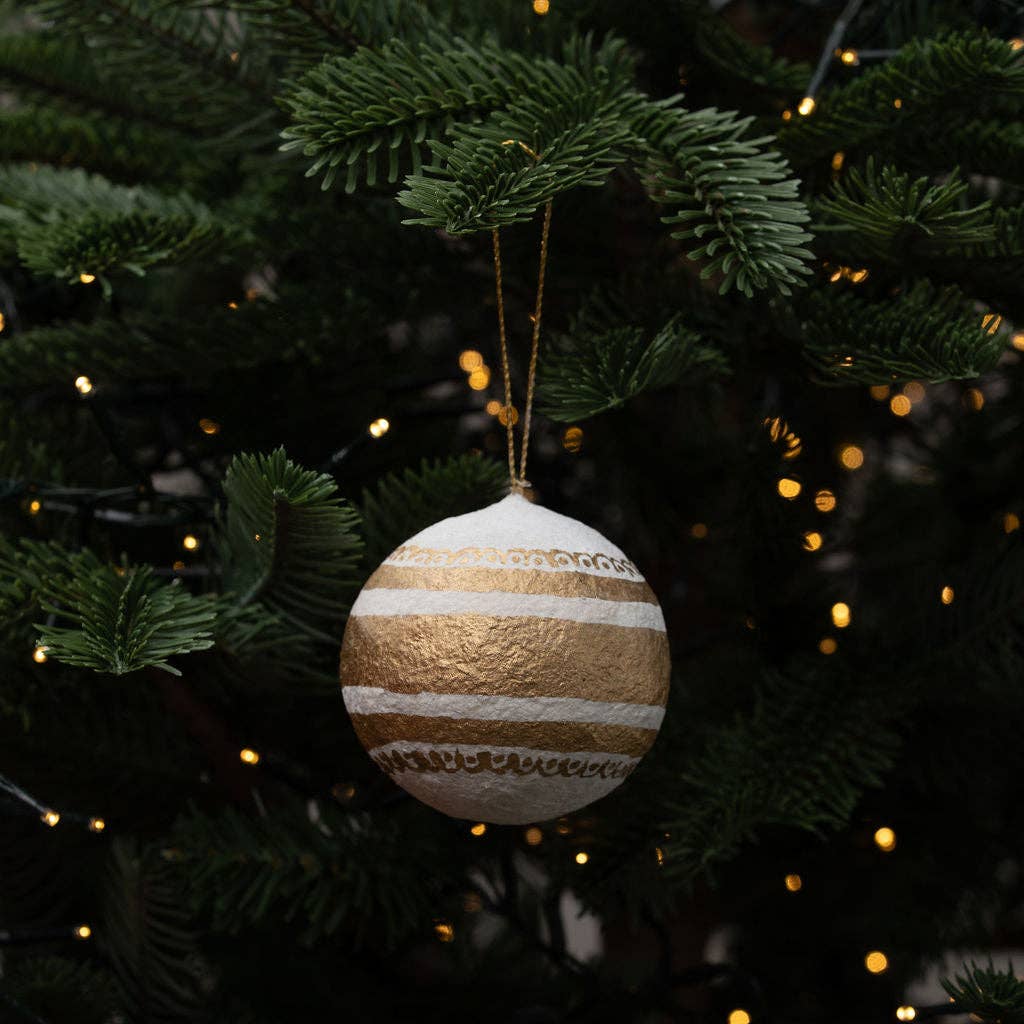 A round Christmas ornament with horizontal gold and white textured stripes hangs from a gold string on a dark green Christmas tree with warm string lights in the background.