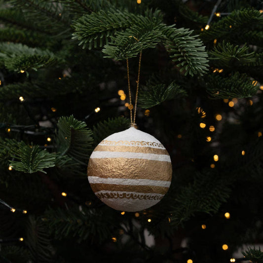 A round Christmas ornament with horizontal gold and white textured stripes hangs from a gold string on a dark green Christmas tree with warm string lights in the background.