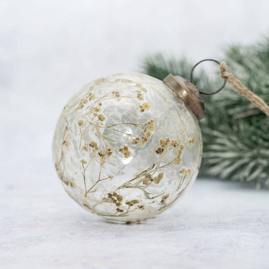A clear glass Christmas ornament decorated with dried botanical elements, featuring a brown metal cap and a hanging twine loop, set against a soft white background with blurred green pine branches.