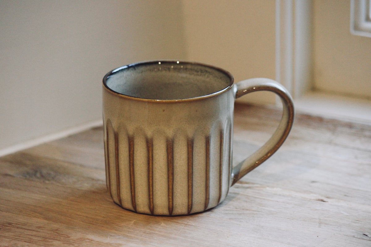 A nkuku ceramic mug with a ribbed vertical texture and a curved handle, sitting on a wooden tabletop in a warm indoor setting.