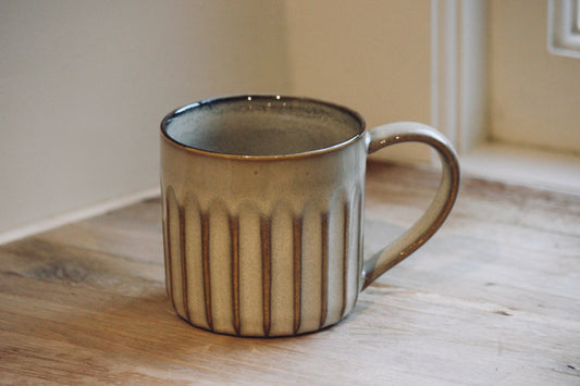 A nkuku ceramic mug with a ribbed vertical texture and a curved handle, sitting on a wooden tabletop in a warm indoor setting.