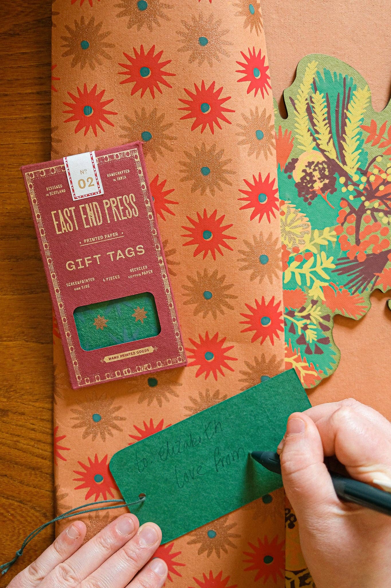Person writing on a green gift tag with a box of East End Press gift tags and floral-patterned paper in the background.