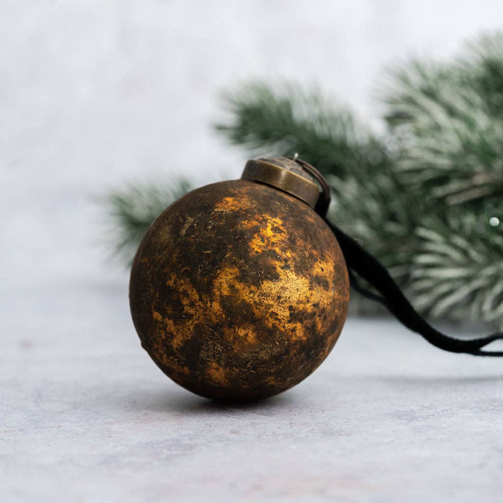 A round distressed metal Christmas ornament with a textured gold and dark brown patina finish, featuring a black hanging ribbon, set against a soft gray background with blurred green pine branches.