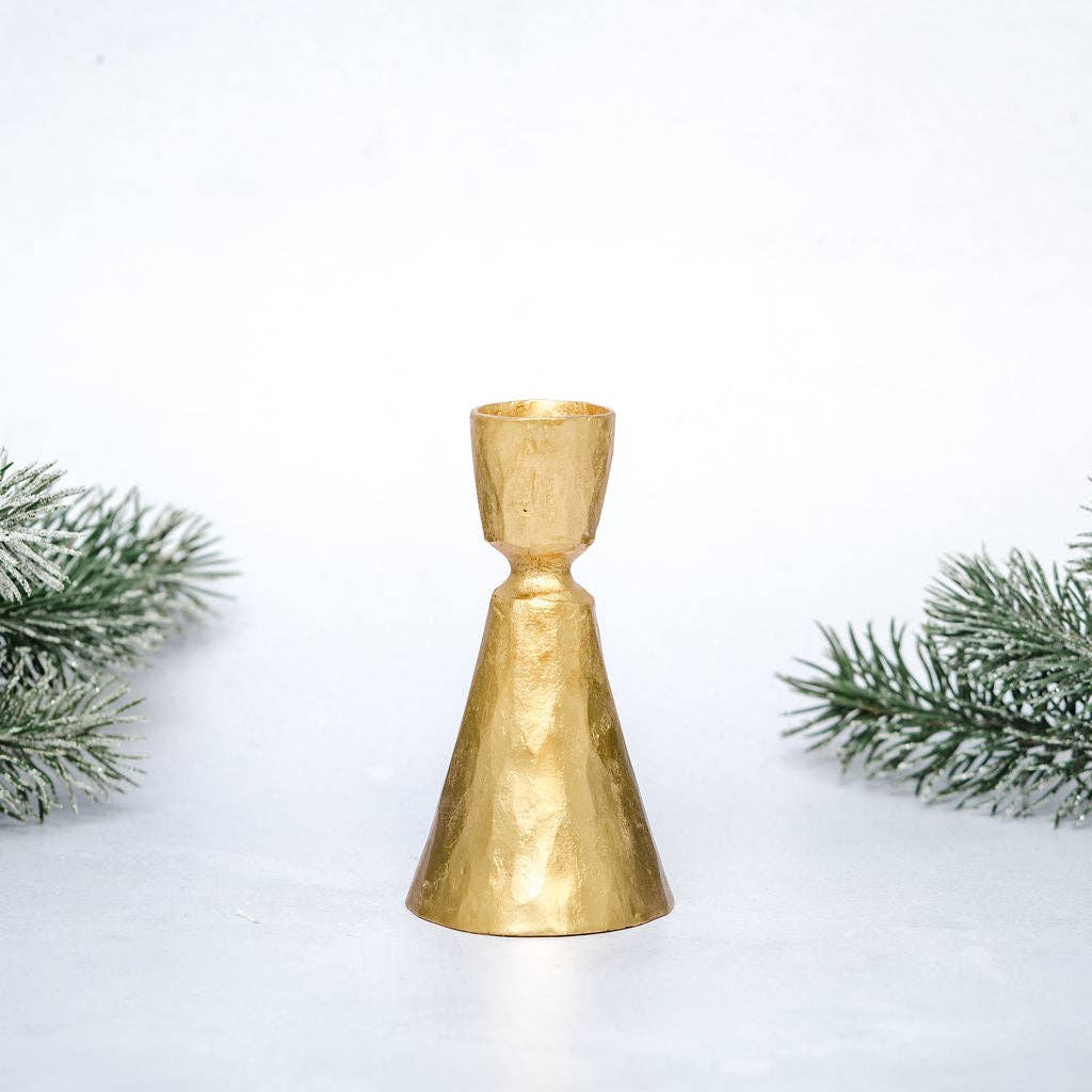 A small metallic gold cone-shaped candle holder or candle stand with a textured, hammered finish, centered on a white surface with decorative green pine branches on both sides.