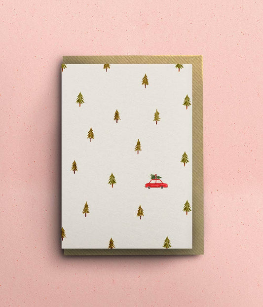 Festive greeting card with a red car and green trees on a white background, against a pink wall.