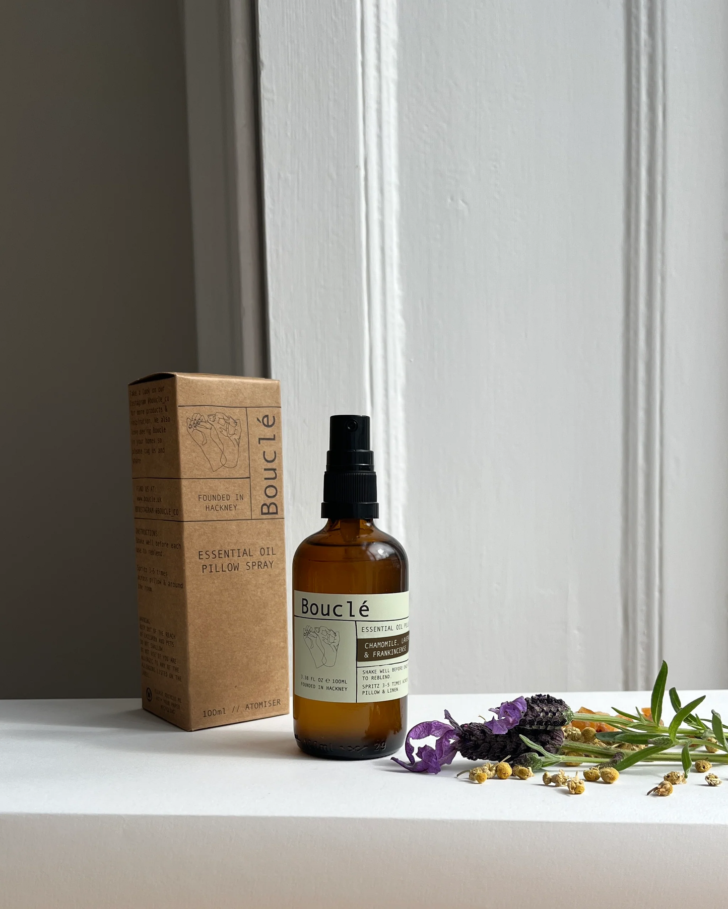 Bottle of Boucle essential oil spray with packaging and herbs on a white surface.