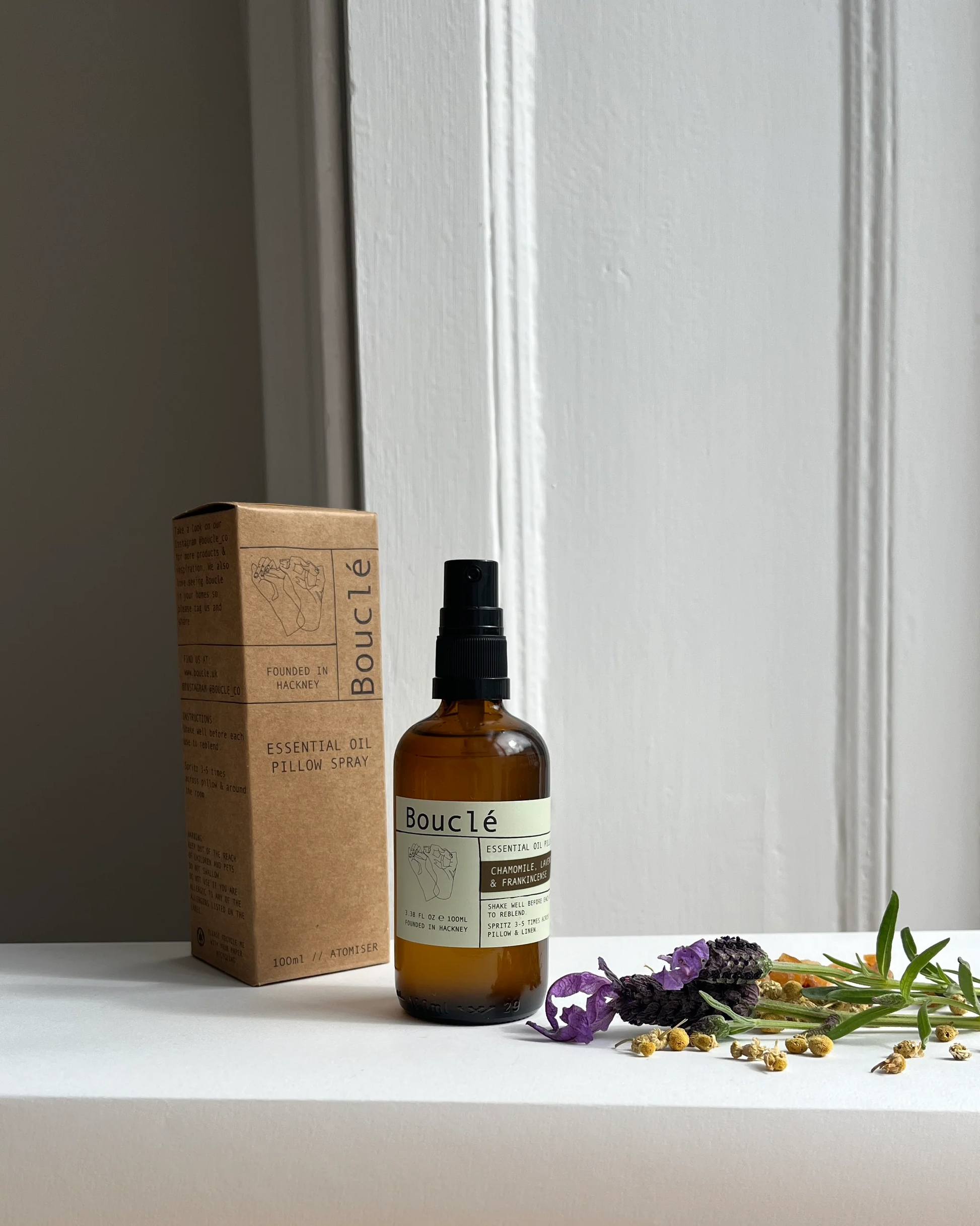 Bottle of Boucle essential oil spray with packaging and herbs on a white surface.
