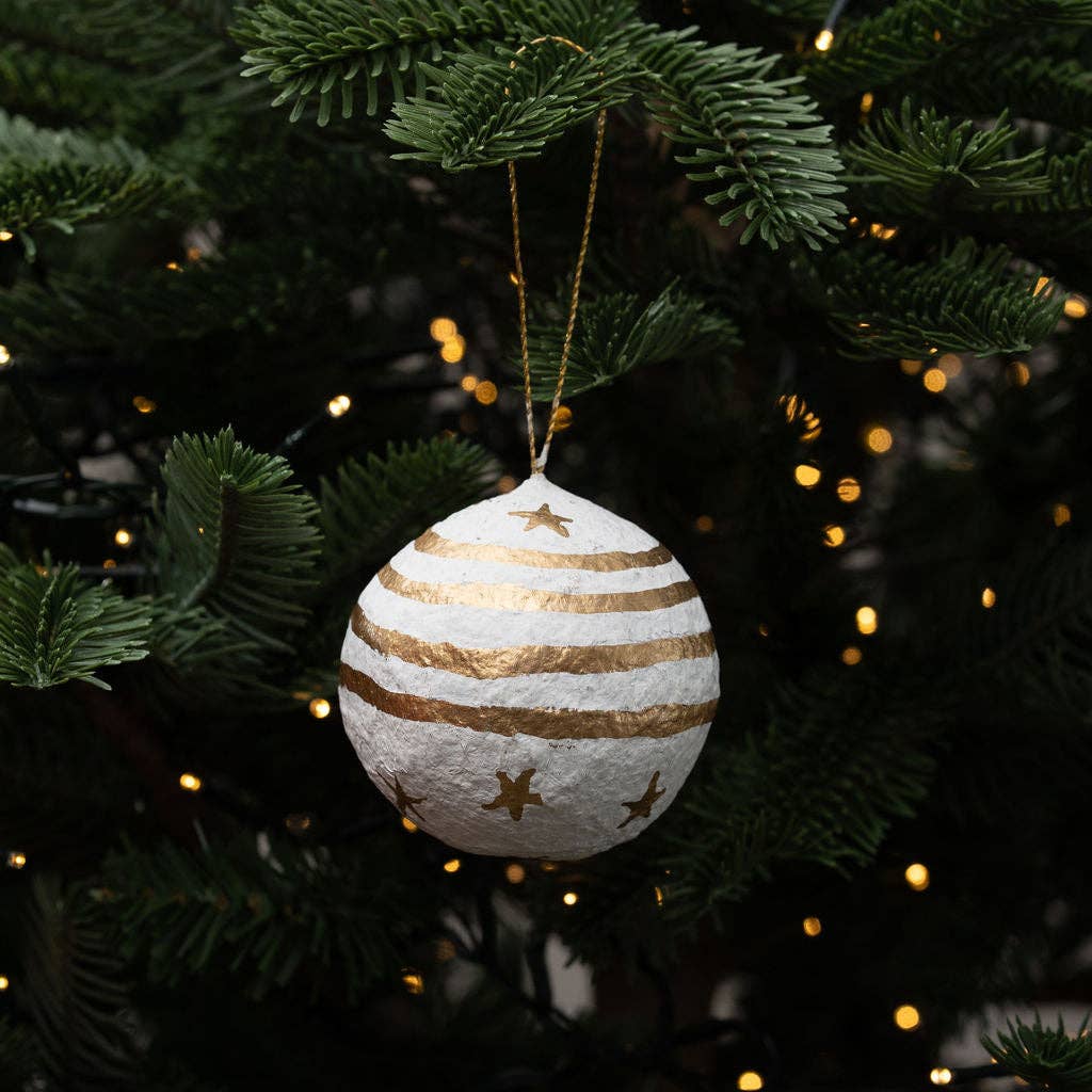 A round white Christmas ornament hand painted using cotton mache and made in India with gold stripes and gold star cutouts, hanging from a gold string on a green Christmas tree with warm lights in the background.