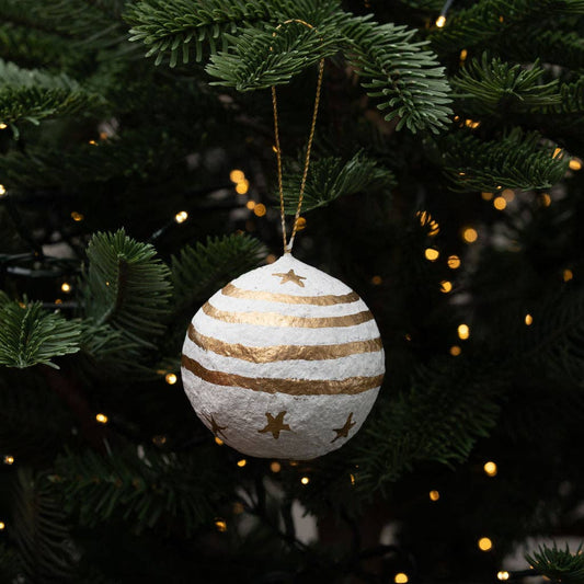 A round white Christmas ornament hand painted using cotton mache and made in India with gold stripes and gold star cutouts, hanging from a gold string on a green Christmas tree with warm lights in the background.