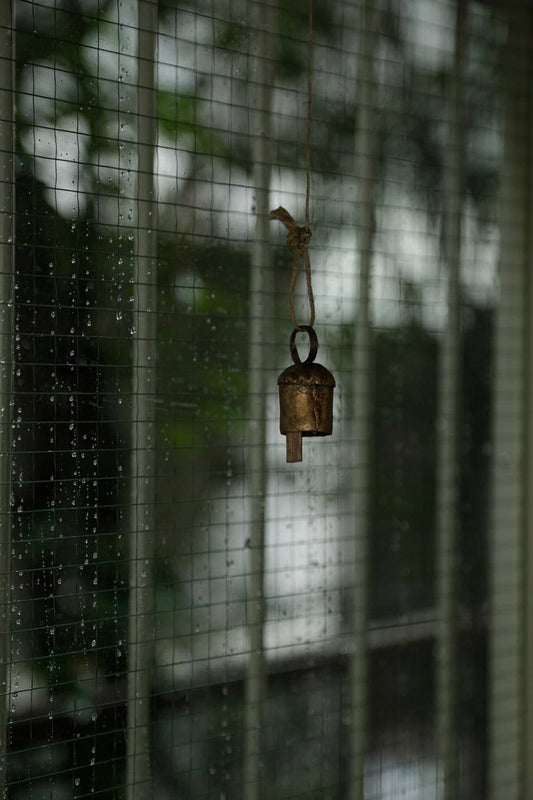A small hanging Noah metal bell with a rope, seen through a window with a grid.