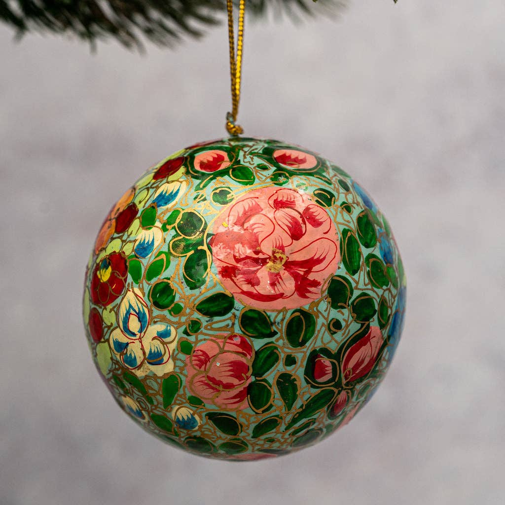 A hand-painted paper mache Christmas ornament featuring pink rose motifs and green leaves on a light blue background, hanging from a gold string under pine branches.