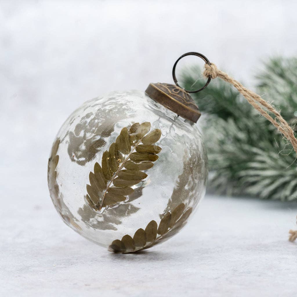 A clear glass Christmas ornament ball decorated with dried olive-green fern leaves, featuring a bronze metal cap and a hanging twine loop, set against a soft, snowy background with blurred greenery.