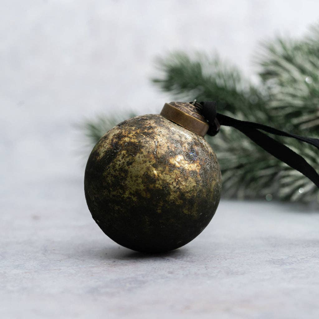 A spherical Christmas ornament with a distressed metallic gold and black finish, featuring a black hanging ribbon, set against a soft gray background with blurred green pine branches.