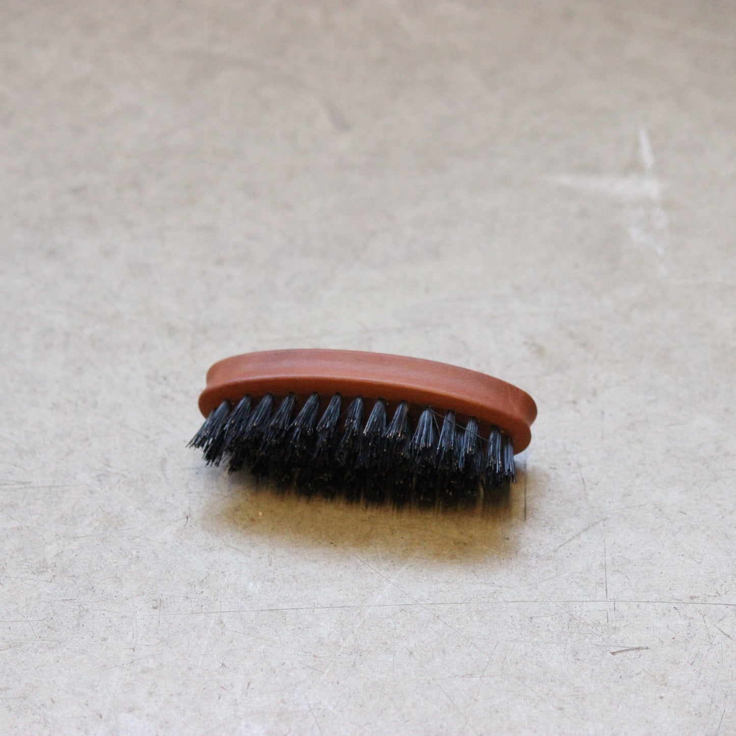 A handcrafted oval wooden beard and moustache brush with dark bristles and a light wooden handle lying on a beige surface.