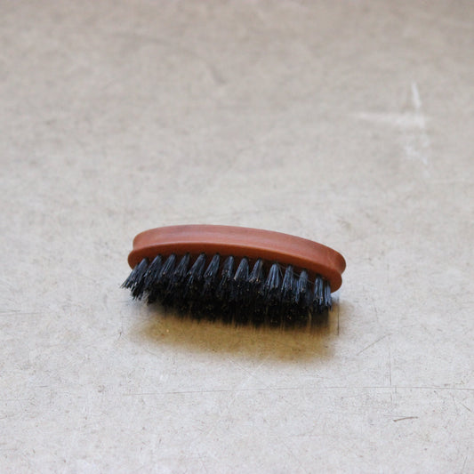A handcrafted oval wooden beard and moustache brush with dark bristles and a light wooden handle lying on a beige surface.