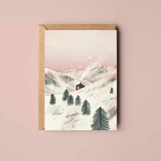 A greeting card with a kraft envelope, featuring an illustration of a snowy mountain landscape with a small cabin and evergreen trees, set against a solid dusty pink background. Snowy Bothy Greetings Card by Rachel Akerman at The Country Crib