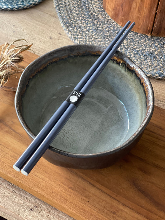 A pair of navy blue chopsticks resting on a rustic ceramic bowl on a wooden table.
