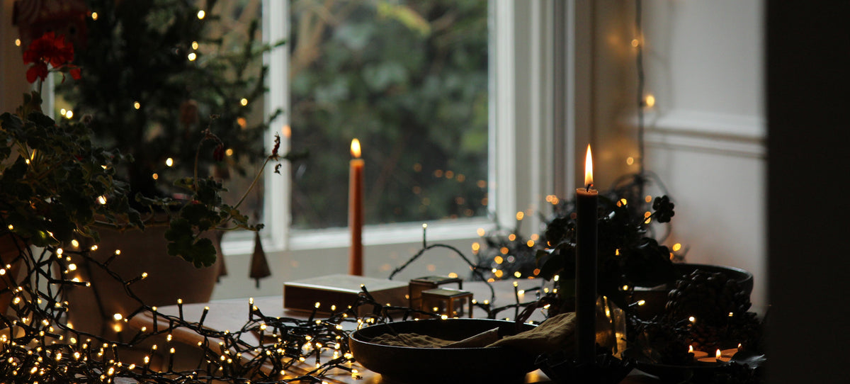 A cozy festive holiday tabletop scene by a window with lit candles, warm string lights, greenery, and wrapped gifts in a softly blurred indoor setting.