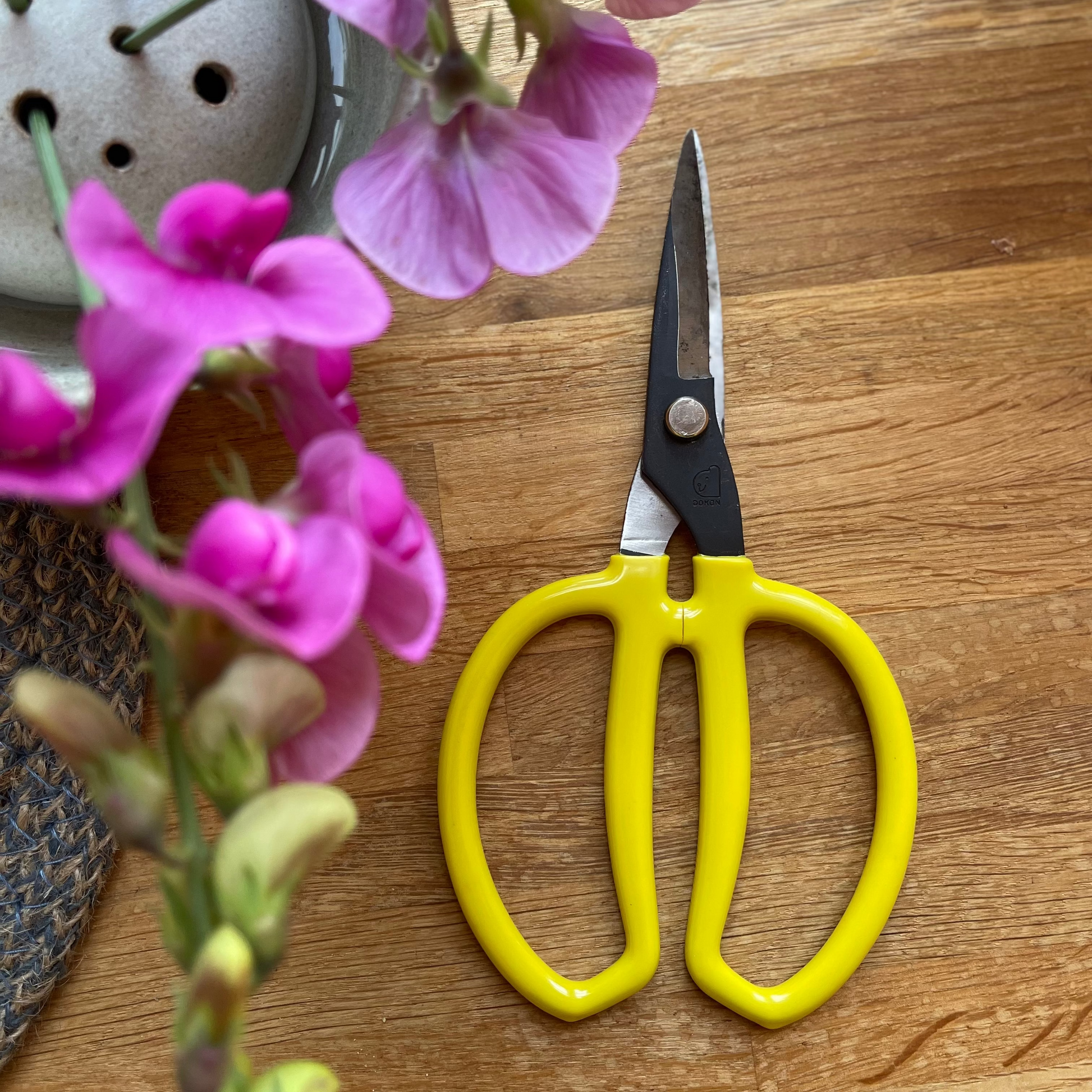 Yellow scissors on a wooden surface with pink flowers and a small pot.