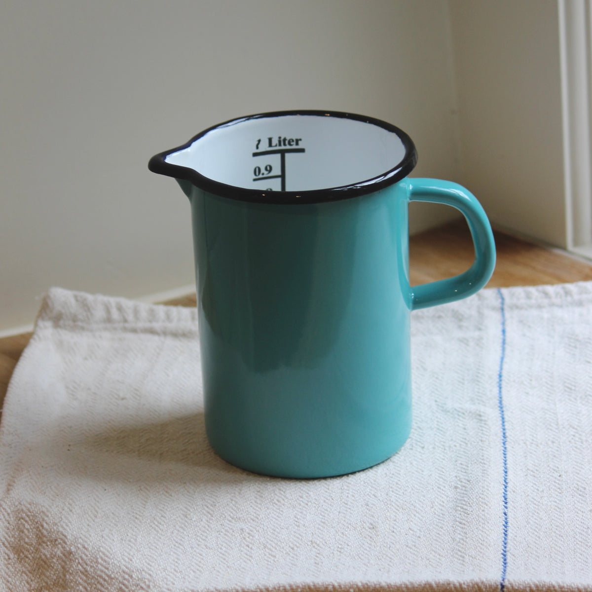 A turquoise enamel measuring pitcher with black rim and handle, sitting on a white kitchen towel with a blue stripe.
