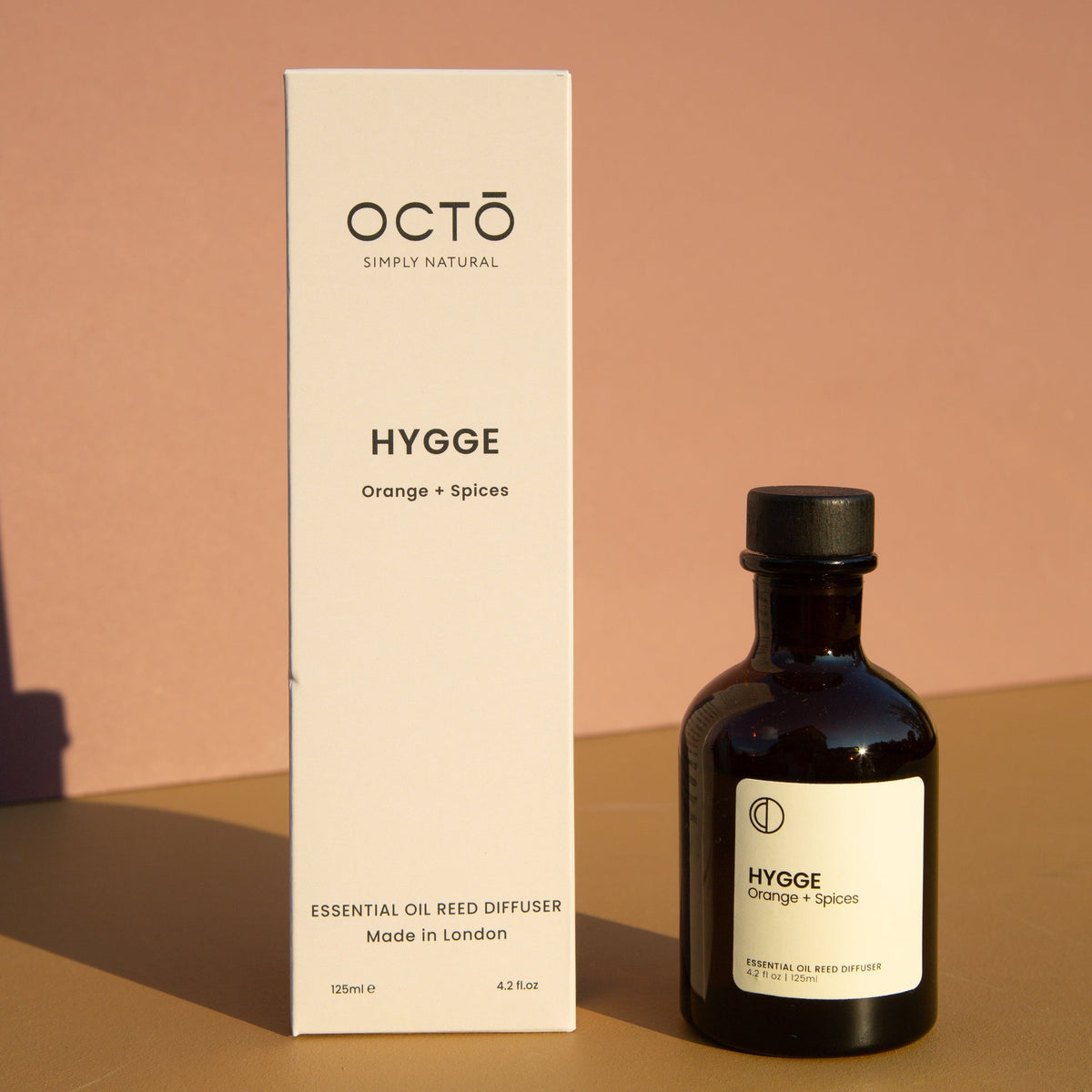 A bottle of OCTØ Hygge essential oil reed diffuser next to its tall white packaging box, set on a warm beige surface against a dusty rose background.