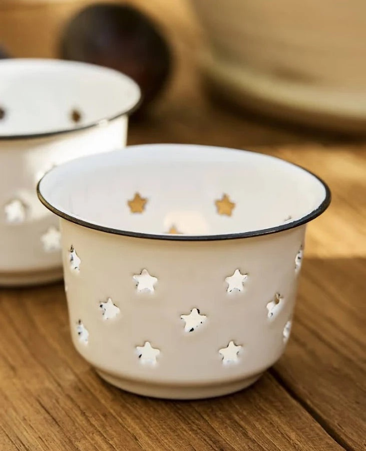 A close-up of a cream-colored enamel candle holder with star-shaped cutouts and a black rim, shown on a wooden surface with another similar holder blurred in the background.
