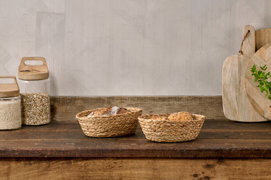 Two woven baskets on a wooden shelf with jars and a cutting board in the background.