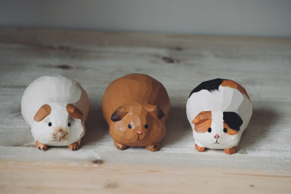 Three hand-painted wooden guinea pig figurines arranged on a light wooden surface against a neutral gray background.