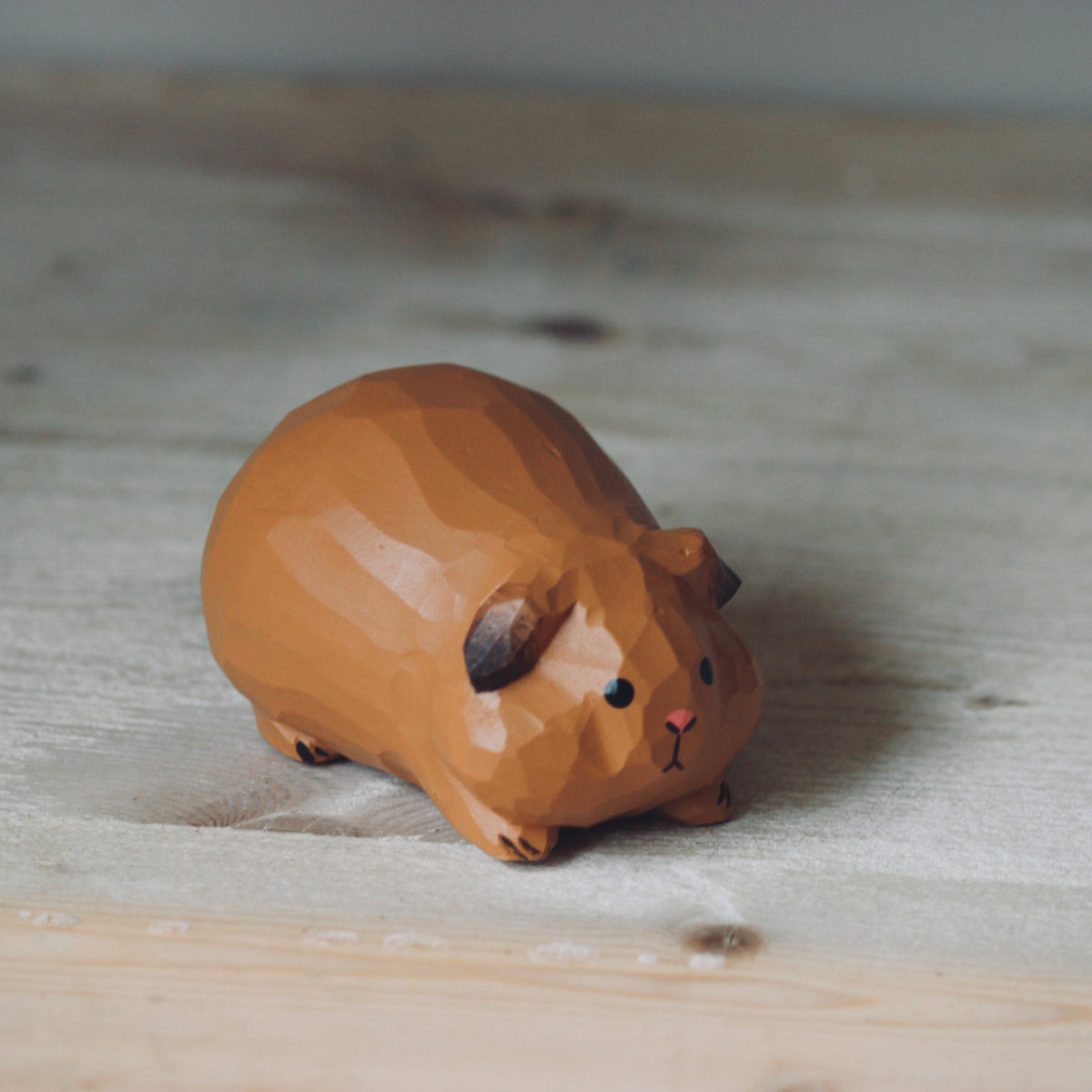 A small style guinea pig figurine in warm brown, resting on a light-colored wooden surface.