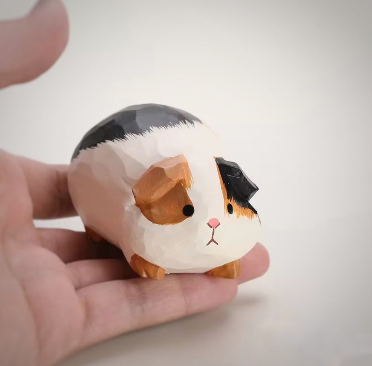 A small hand-painted guinea pig figurine resting on a person's palm, showcasing its triangular facets and realistic painted details.