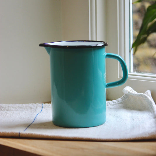 A turquoise enamel pitcher with a black rim sits on a white kitchen towel on a wooden windowsill.