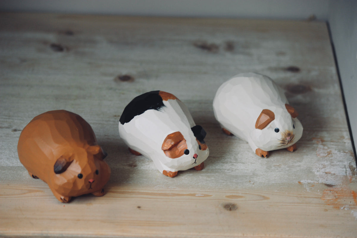 Three small painted guinea pig figurines arranged on a light wood surface. One figurine is solid brown, while the other two are mostly white with black and brown painted details.