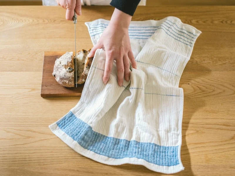 Person using a bread knife on a wooden cutting board with a towel underneath.